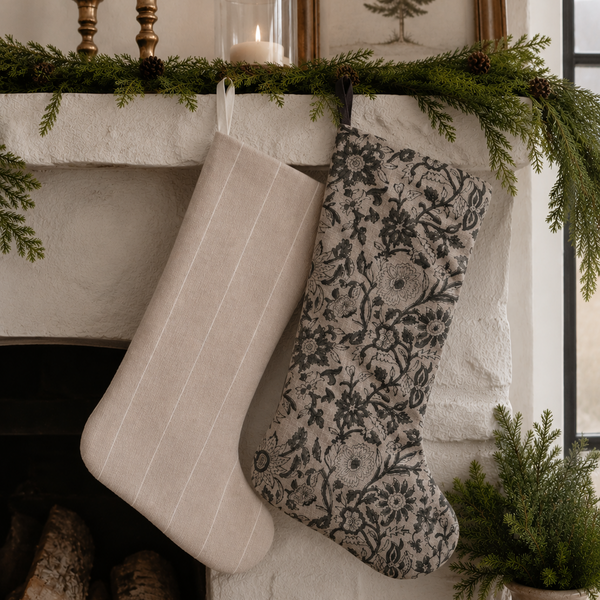 Two handcrafted Christmas stockings in neutral linen and vintage floral designer fabric hanging on a fireplace mantel with evergreen garland in a luxury holiday home.