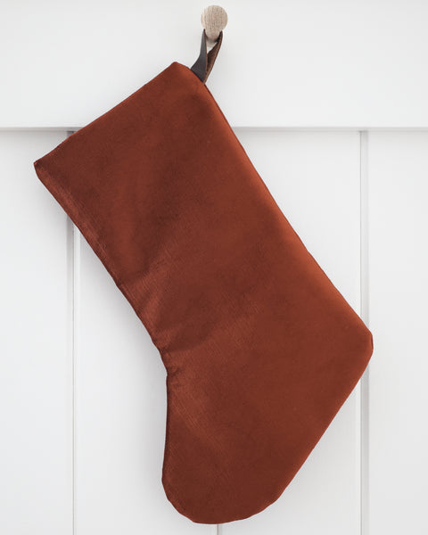 Cider Velvet Stocking, Boho Design Christmas stocking in rich rust-terracotta polyester velvet with a dark brown strap hook, hanging from a wooden peg on a white paneled wall