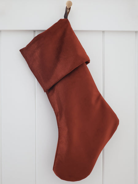 Cider Velvet Stocking, Heritage Design cuffed Christmas stocking in rich rust-terracotta polyester velvet with a dark brown strap hook, hanging from a wooden peg on a white paneled wall