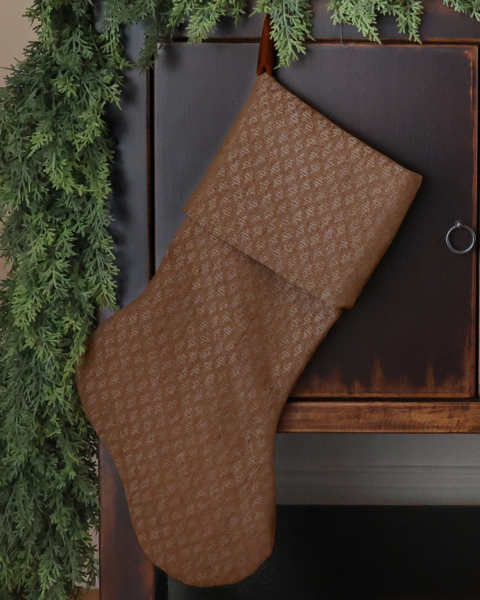 HACKNER HOME handmade golden brown textured Christmas stocking in Heritage cuff design, featuring richly woven designer fabric and warm neutral tones, styled against dark wood with evergreen garland.