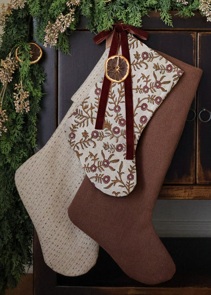 HACKNER HOME luxury handmade Christmas stockings in textured ivory, vintage floral linen, and warm red-brown designer fabrics, styled with burgundy velvet ribbon, dried orange, and evergreen garland.