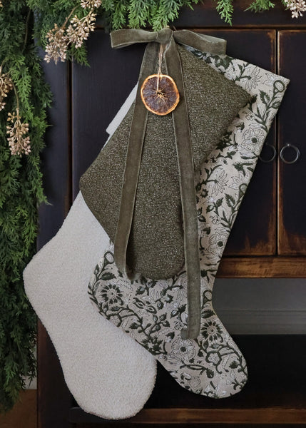 HACKNER HOME luxury handmade Christmas stockings in dark olive green textured woven fabric, green floral print, and ivory boucle, styled with velvet ribbon and evergreen garland.