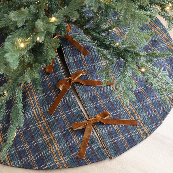 Blue tartan plaid handmade Christmas tree skirt with green and golden stripes and brown velvet ribbon ties beneath a Christmas tree.