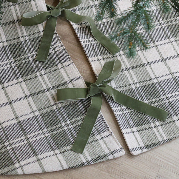 Sage green and ivory plaid handmade Christmas tree skirt with green velvet ribbon bows beneath an evergreen Christmas tree close up.