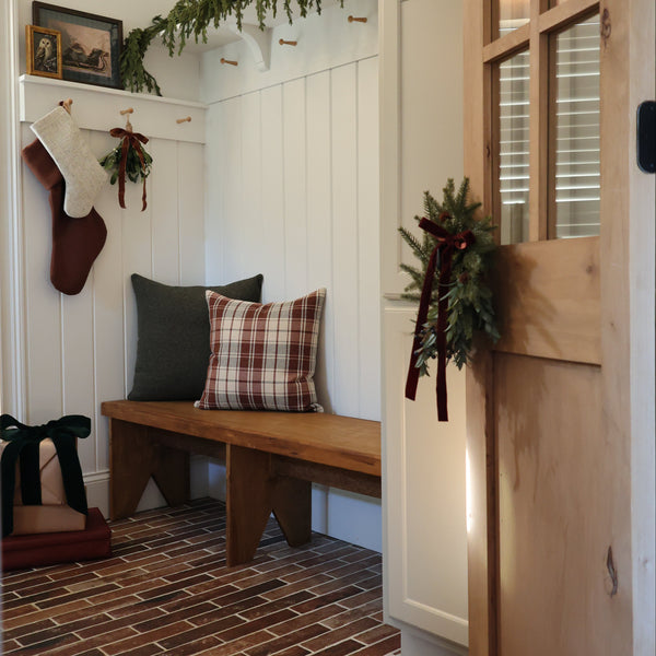 HACKNER HOME handmade red-brown plaid pillow cover layered with a forest green pillow on a wooden entryway bench, styled with Christmas stockings, greenery, and warm natural wood accents.
