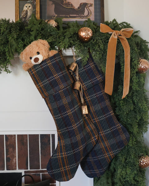 Two HACKNER HOME handmade blue tartan plaid Christmas stockings with navy, forest green, and golden brown tones, styled with a teddy bear, evergreen garland, velvet ribbon, and brass bells.