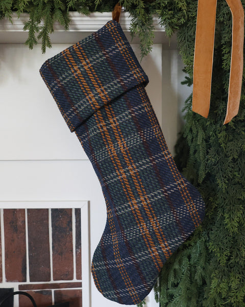 HACKNER HOME handmade blue tartan plaid Christmas stocking in Heritage cuff design, featuring navy blue, forest green, and golden brown woven plaid fabric, styled with evergreen garland.