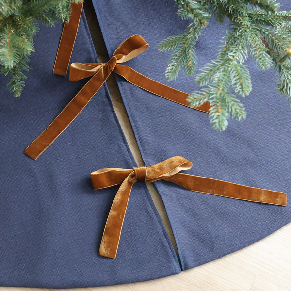 Navy blue handmade Christmas tree skirt with brown velvet ribbon bows beneath an evergreen Christmas tree.
