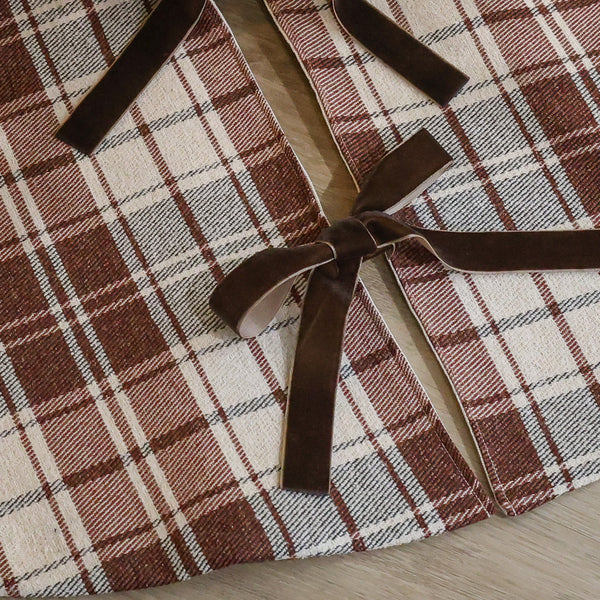 Handmade red-brown plaid Christmas tree skirt with warm cream background and brown velvet ribbon ties beneath a Christmas tree