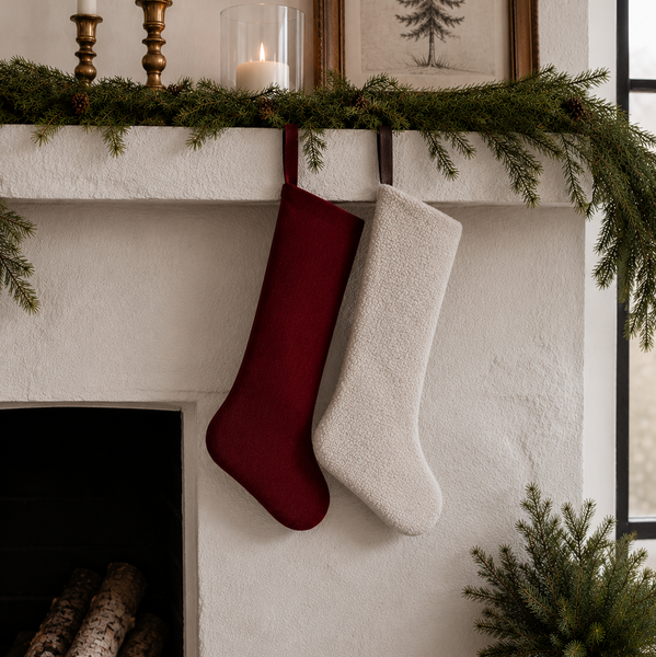 Two Christmas stockings hang from a white fireplace mantel decorated with green garland, candles, and framed art. One stocking is deep burgundy and the other is cream white, with logs visible in the fireplace below.