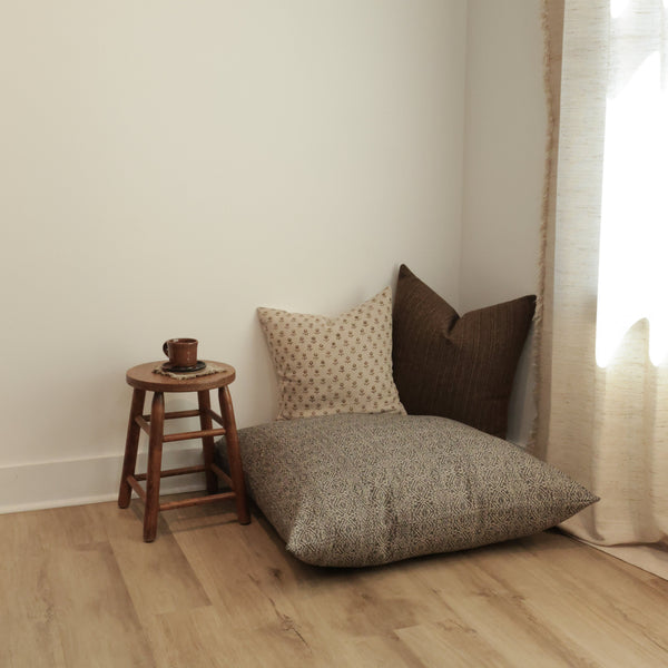 “Cozy styled corner featuring the Hackner Home Acadia floor pillow cover layered with two accent pillows beside a rustic wooden stool holding a mug, set against a light wall and soft natural lighting.