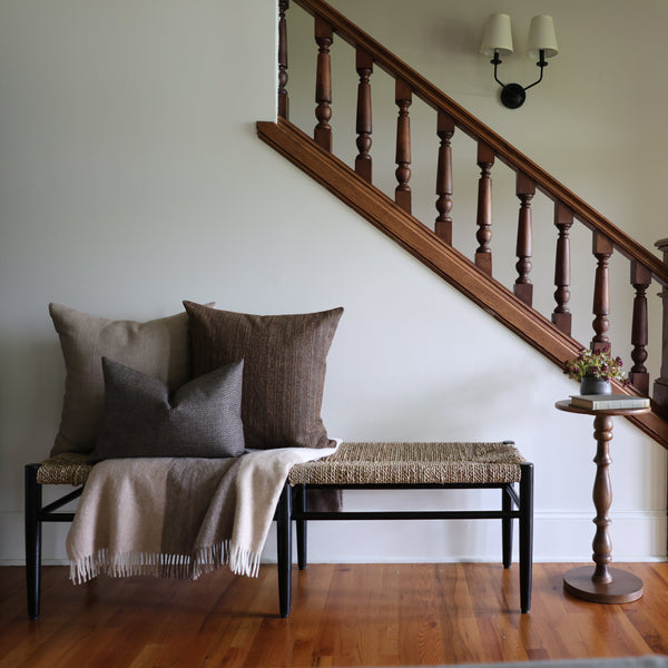 Entryway bench styled with neutral and brown throw pillows and a beige fringed wool blanket, beside a small wooden side table and staircase with wooden railing.