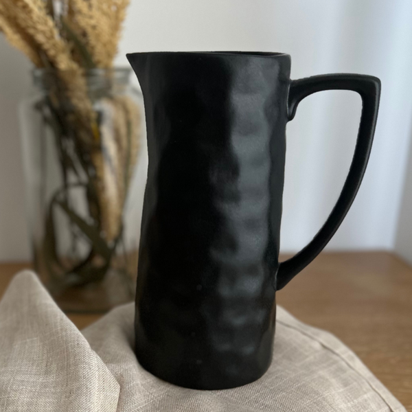 Hackner Home black ceramic serving pitcher with a hand-formed textured finish displayed on a neutral linen cloth with dried floral decor in the background.