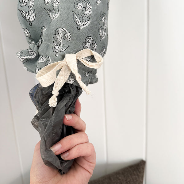 Hand pulling a plastic bag from the bottom of Hackner Home blue linen grocery bag holder with white floral pattern and cream drawstring tie.