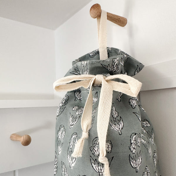 Close-up of Hackner Home blue linen grocery bag holder with white floral print and cream drawstring tie, hanging on a wooden peg in a modern farmhouse laundry room.
