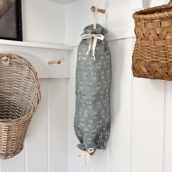 Hackner Home blue linen grocery bag holder with white floral pattern, hanging in a farmhouse-style laundry room with woven baskets and wooden peg hooks.