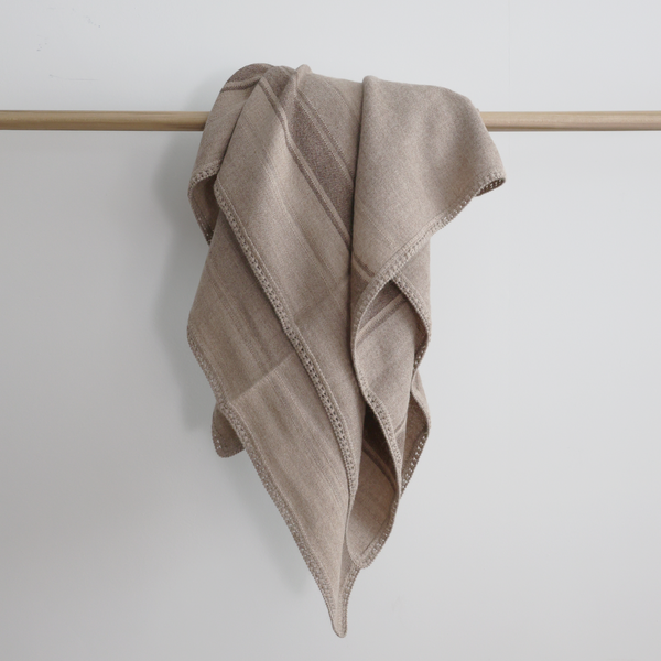 Soft woven baby alpaca throw blanket in light brown tones with subtle stripes and delicate crocheted edge, draped over a wooden rod.