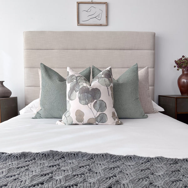 Neutral bedroom featuring Hackner Home Eucalyptus Leaf pillow cover layered with green and beige pillows on a modern upholstered headboard with a gray knit blanket at the foot of the bed.
