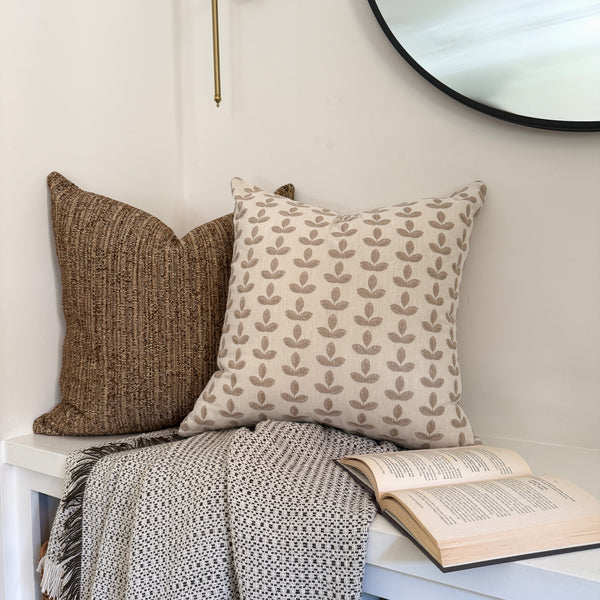Cozy reading nook with two decorative pillows — one brown textured and one cream with tan embroidered leaves — arranged on a white bench with a patterned throw blanket and an open book beneath a round wall mirror.