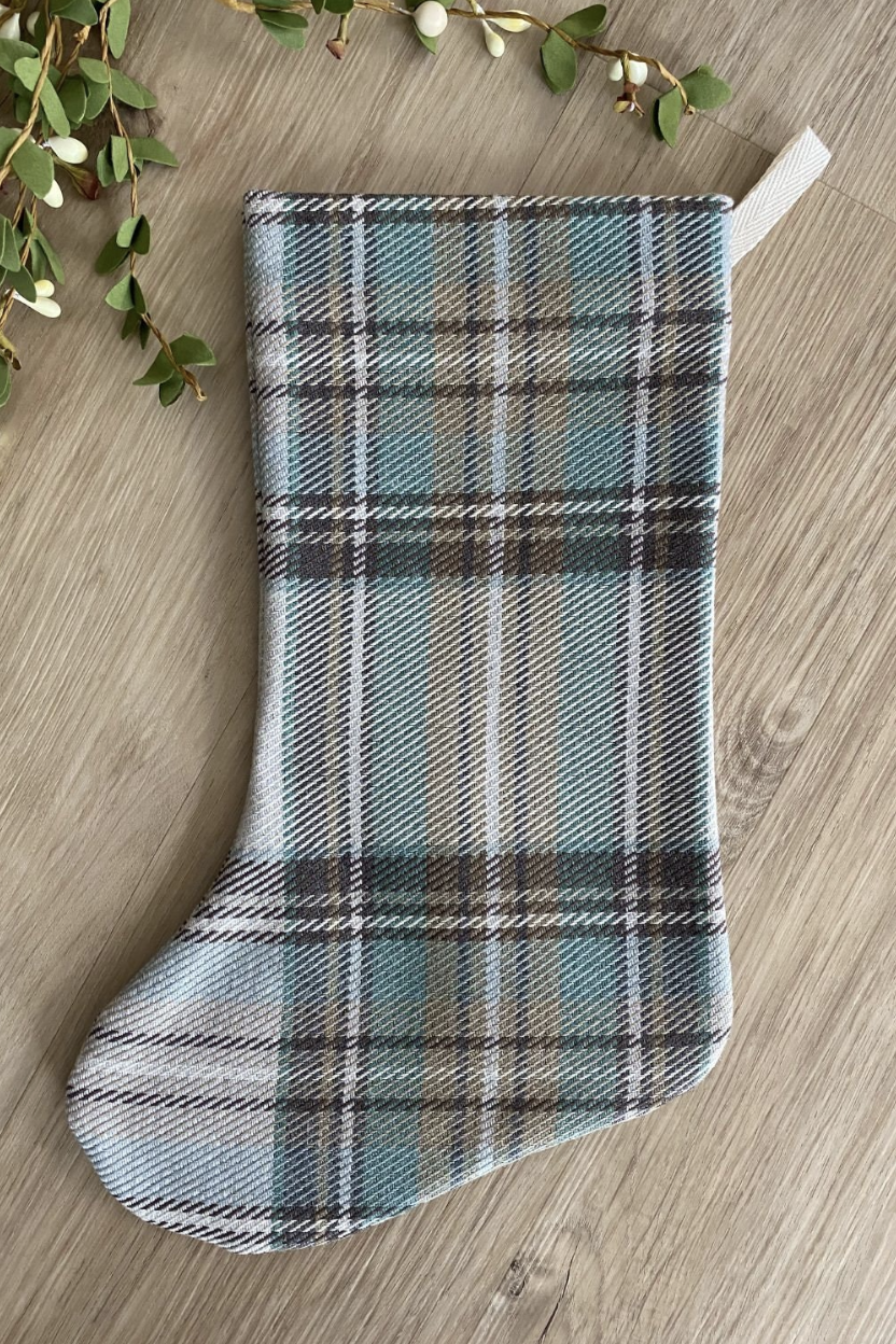 Hackner Home Green Plaid Boho Christmas Stocking featuring a soft woven fabric in sage green, beige, and brown plaid pattern, styled flat on a wood surface with greenery accents.