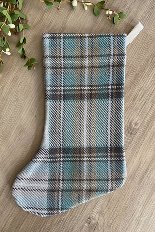 Hackner Home Green Plaid Boho Christmas Stocking featuring a soft woven fabric in sage green, beige, and brown plaid pattern, styled flat on a wood surface with greenery accents.