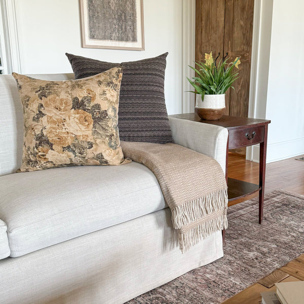 Living room with a light beige sofa styled with a beige fringed throw blanket and two decorative pillows, one floral and one striped, beside a wooden side table with a potted plant.
