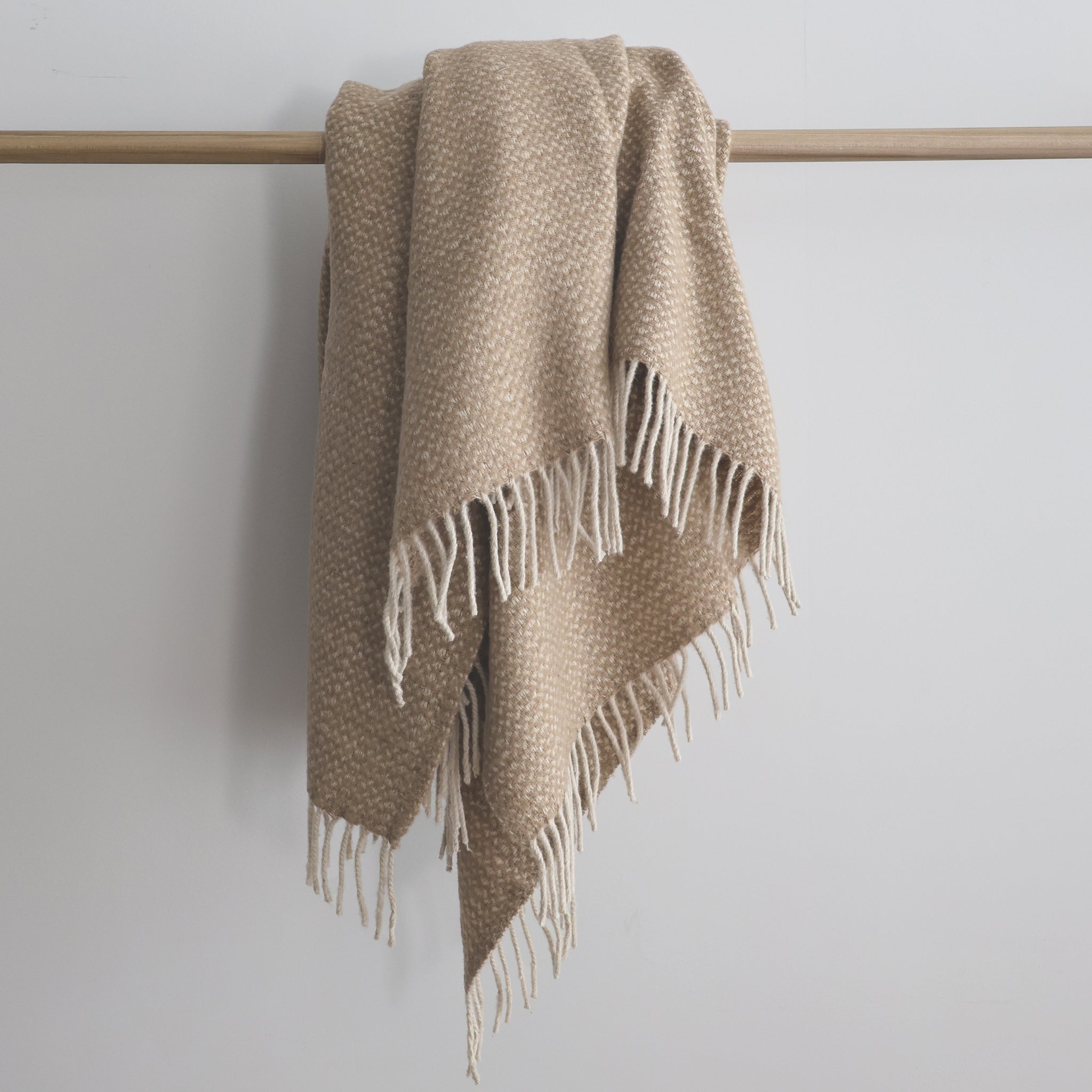 Beige recycled wool blend throw blanket with a subtle woven pattern and cream fringe, draped over a wooden rod.