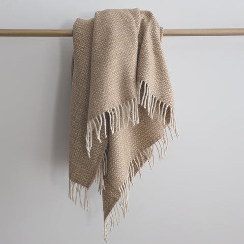 Beige recycled wool blend throw blanket with a subtle woven pattern and cream fringe, draped over a wooden rod.