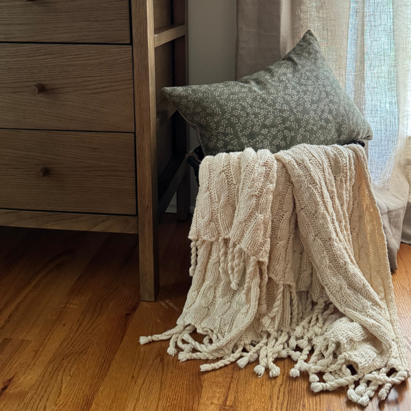 Styled view of Hackner Home Sage Green Floral Block Print pillow cover on a chair with a cream knit throw blanket beside a wooden dresser and linen curtains.