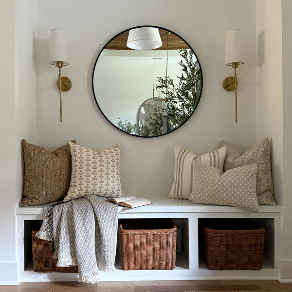 Cozy entryway bench styled with neutral and soft sage decorative pillows, a textured throw blanket, woven baskets below, and a round mirror with brass sconces above.